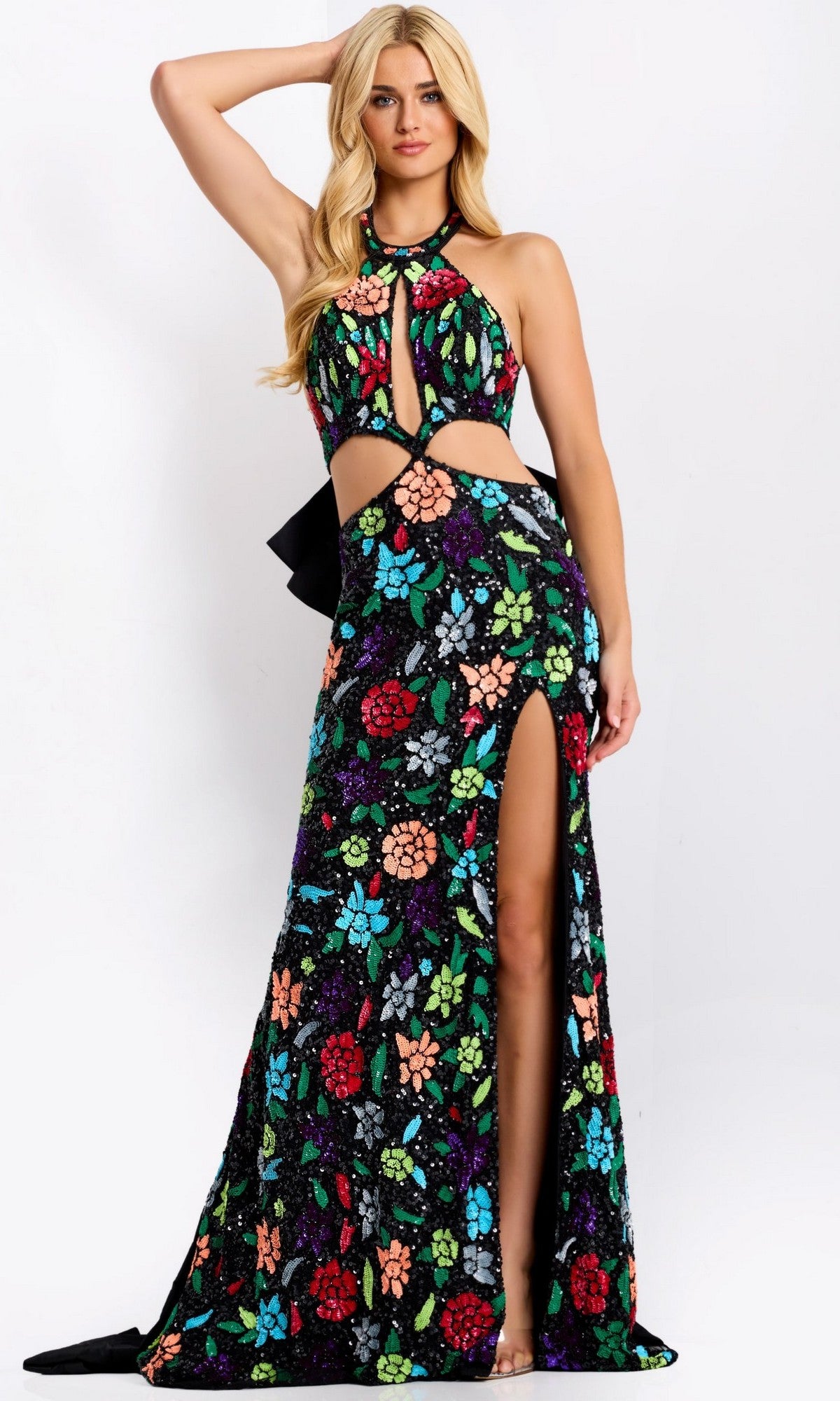 A woman wears the Jovani 49251 long prom dress—a sleeveless, floor-length black gown with a halter neckline, vibrant floral patterns, waist cutouts, and a thigh-high slit—standing against a plain background.